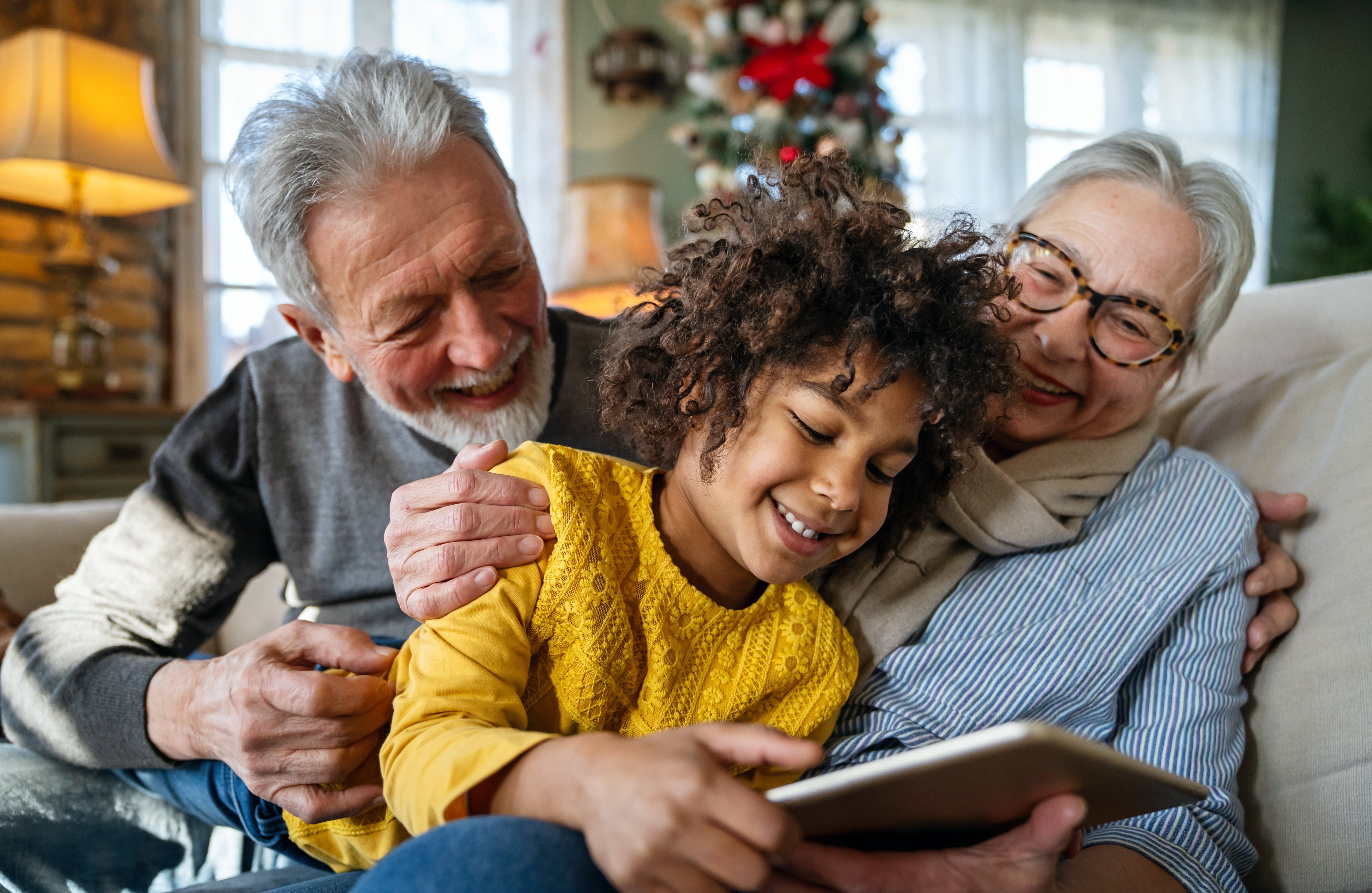 A family of three is sitting on a couch with a tablet in the middle person's hands.